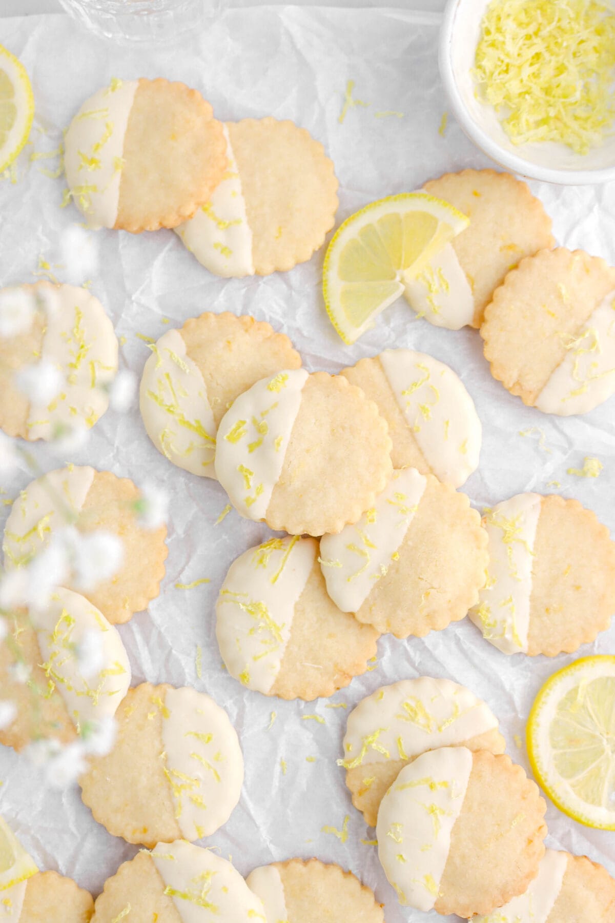 overhead shot of lemon shortbread cookies with bowl of lemon zest, lemon slices around and white flowers above cookies.