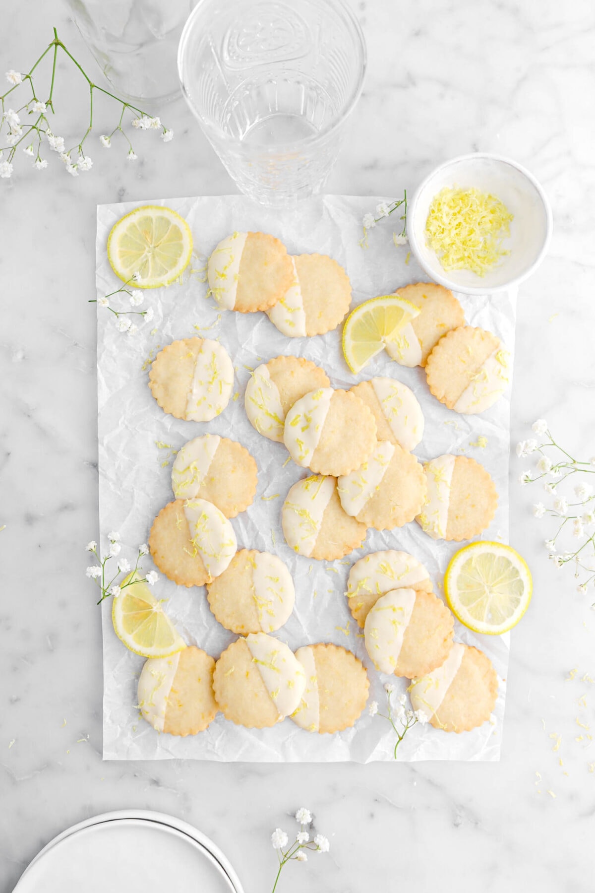 pulled back overhead shot of lemon shortbread cookies on parchment paper with white flowers around, lemon slices, an empty glass, and two small white plates around on marble surface.