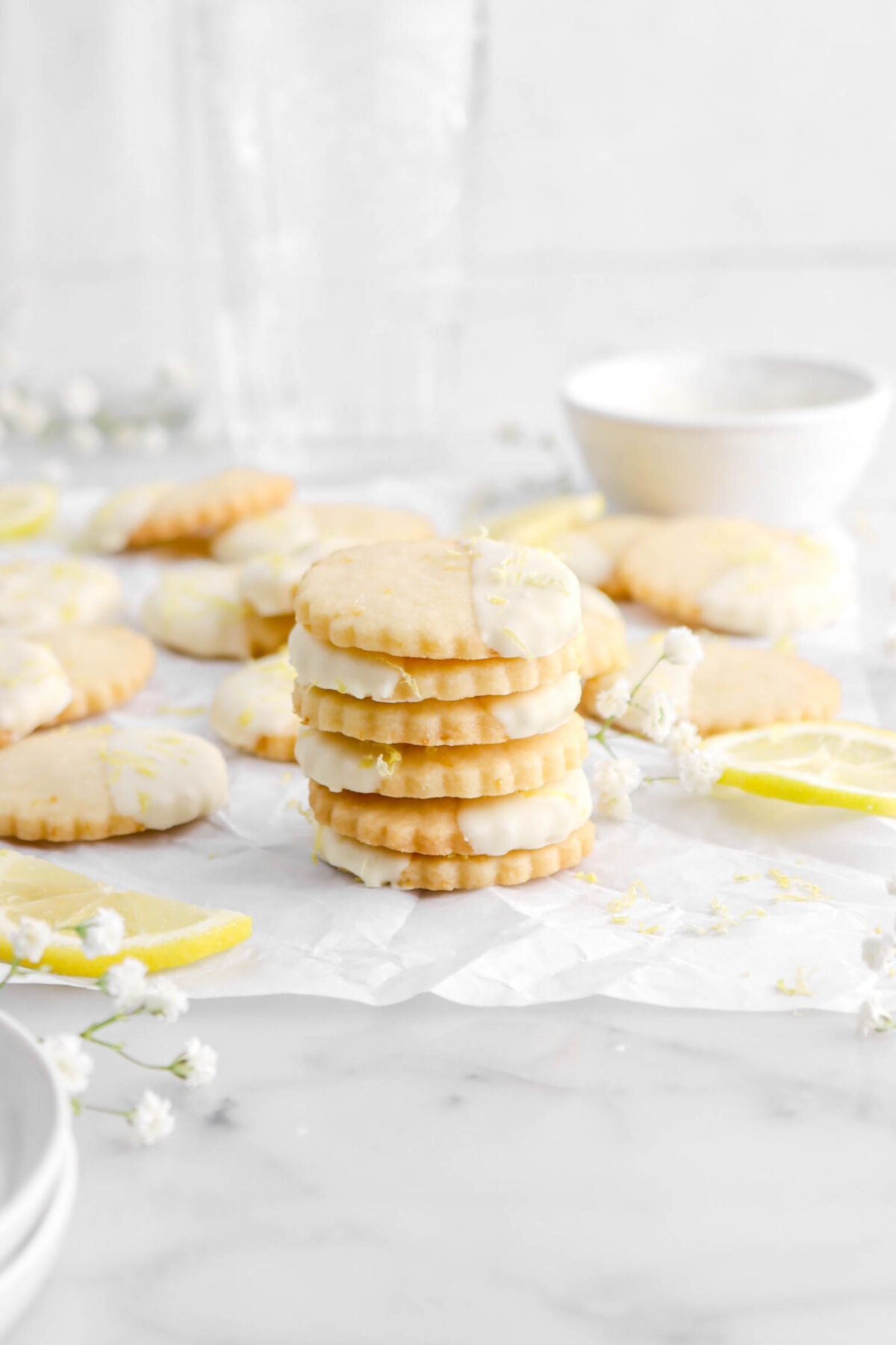 angled shot of six stacked cookies on parchment paper with white flowers and lemon slices around, more cookies behind, a small white bowl, and two empty glasses.