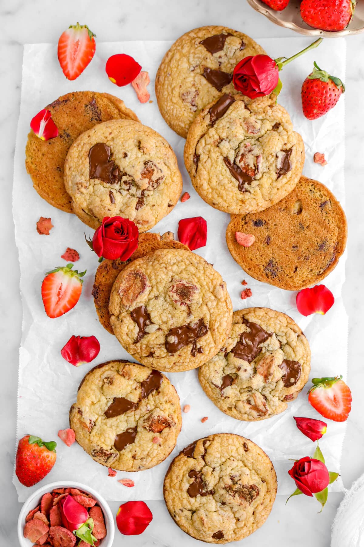 ten chocolate chip strawberry cookies on parchment paper with strawberries, roses, and rose petals around on marble surface with white bowl of freeze dried strawberries beside.