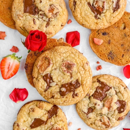 ten strawberry chocolate chip cookies on parchment paper with a rose leaning against cookie and rose petals around.