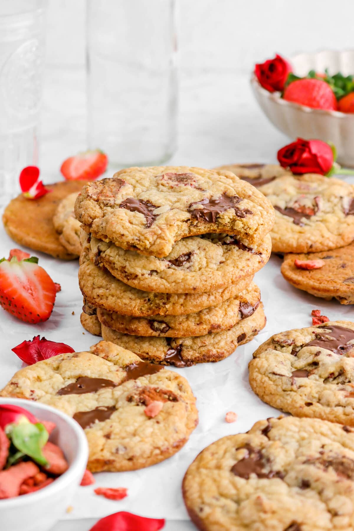 stacked strawberry chocolate chip cookies on parchment paper with more cookies around and bowl of strawberries behind with red roses.
