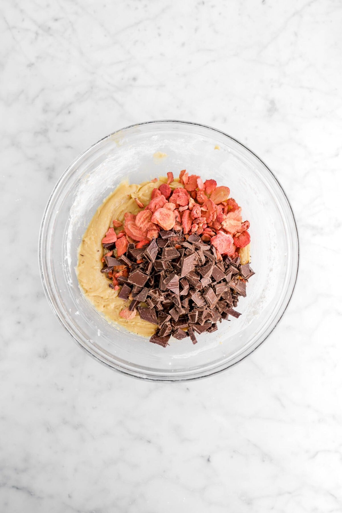 chopped chocolate and freeze-dried strawberries added on top of cookie dough in glass bowl.