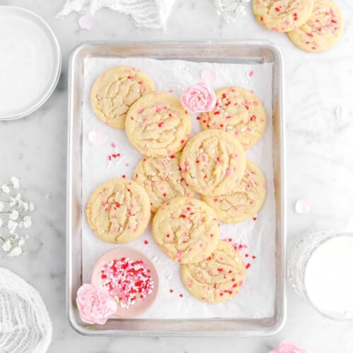 sugar cookies piled on a small parchment lined sheet pan with pink bowl full of valentine's-themed sprinkles, and pink carnations. Three cookies on marble surface around sheet pan with more flowers, a white cheesecloth, a glass of milk, another pink carnation, and white flowers around sheet pan.