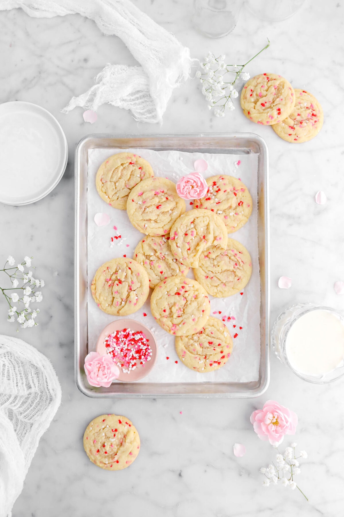 overhead shot of nine sugar cookies in parchment lined sheet pan with one missing a bite, two pink carnations, and pink bowl of sprinkles, with three more cookies around on marble surface, another pink carnation, white flowers, a glass of milk, a stack of plates, and a white cheesecloth beside.