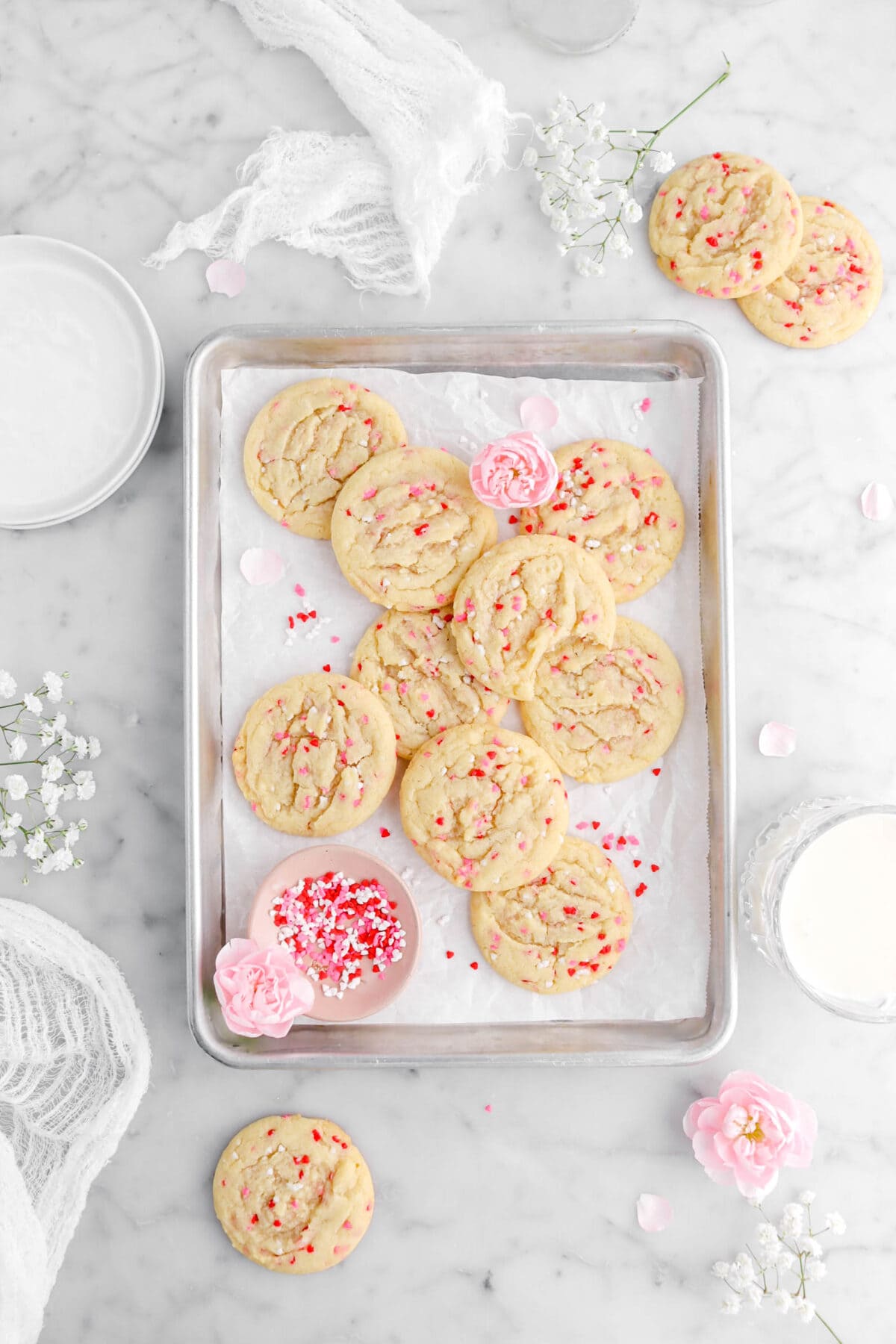 overhead shot of piled sugar cokies in parchment lined sheet pan with one cookie missing a bite, a bowl of sprinkles, and pink carnations in sheet pan, with more cookies and flowers around on marble surface.