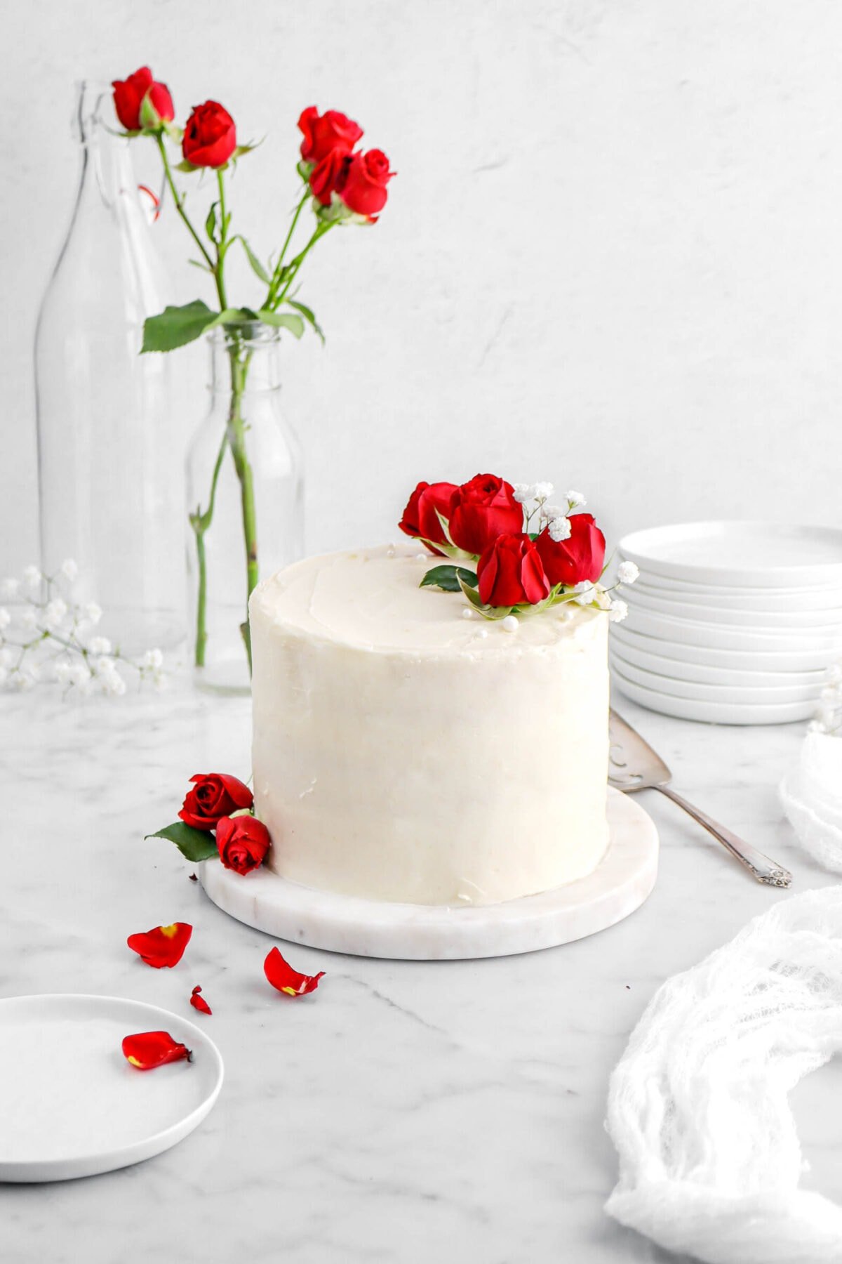 cake on marble board with red roses, small white flowers, and white pearl sugar on top of cake with a plate and rose petals beside, roses behind in glass, and a white cheesecloth beside.
