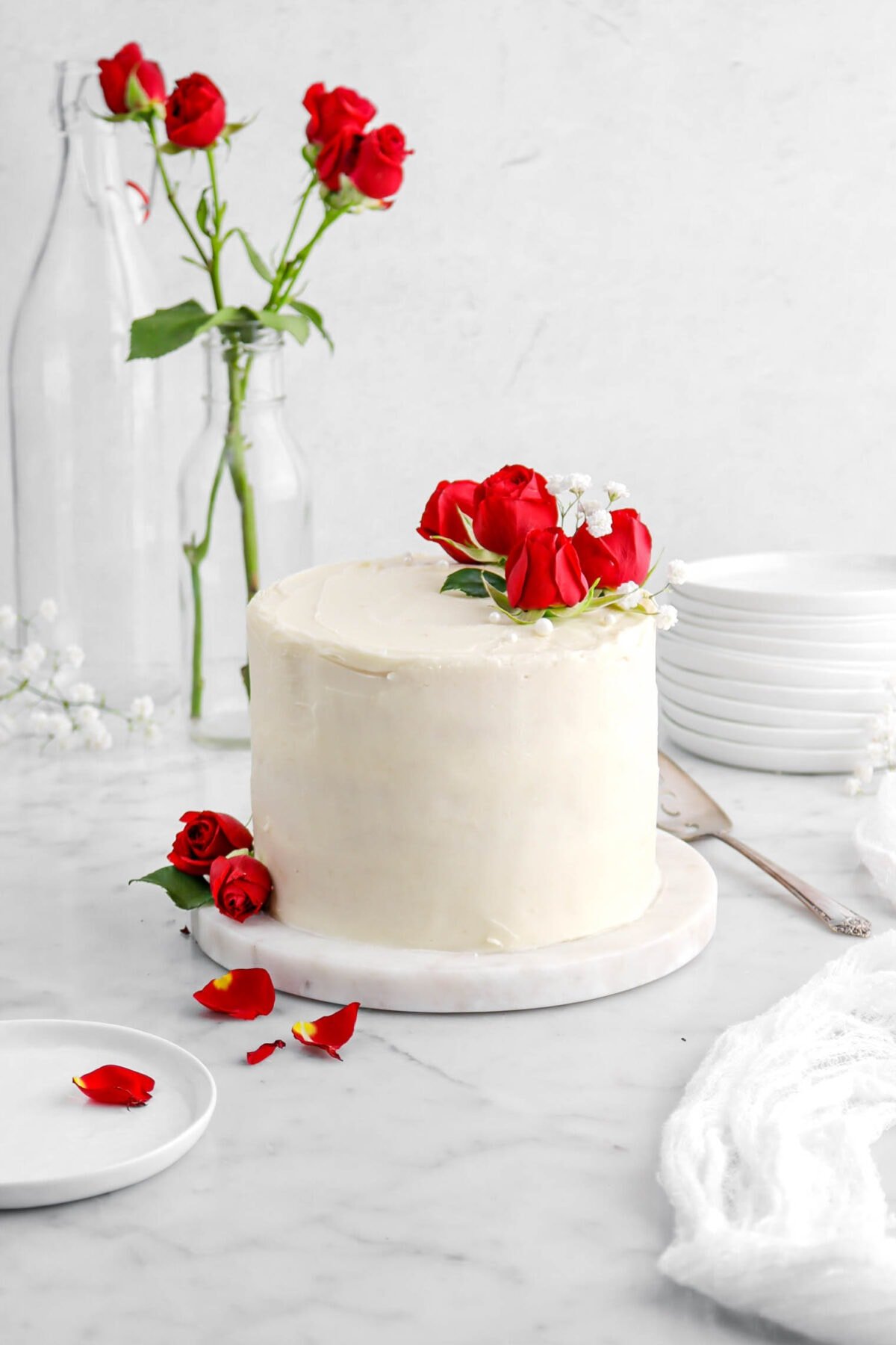 front shot of vanilla rose cake on marble board with roses and white flowers on top, a plate with a rose petal beside, stack of plates, and red roses in a glass behind.