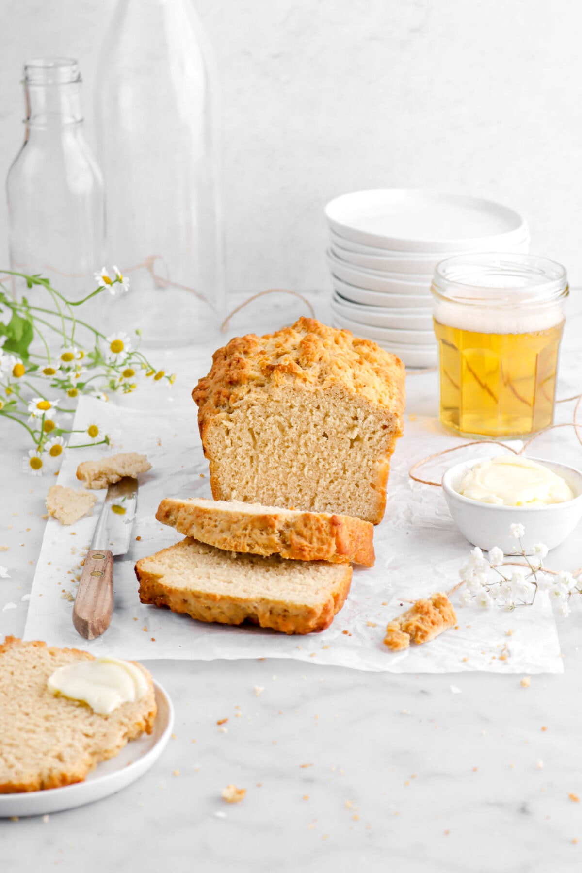 beer bread with two slices laying in front of loaf on parchment paper with bowl of butter and a knife beside, bread crumbs, and flowers around, with another slice of bread on white plate with smear of butter on top.
