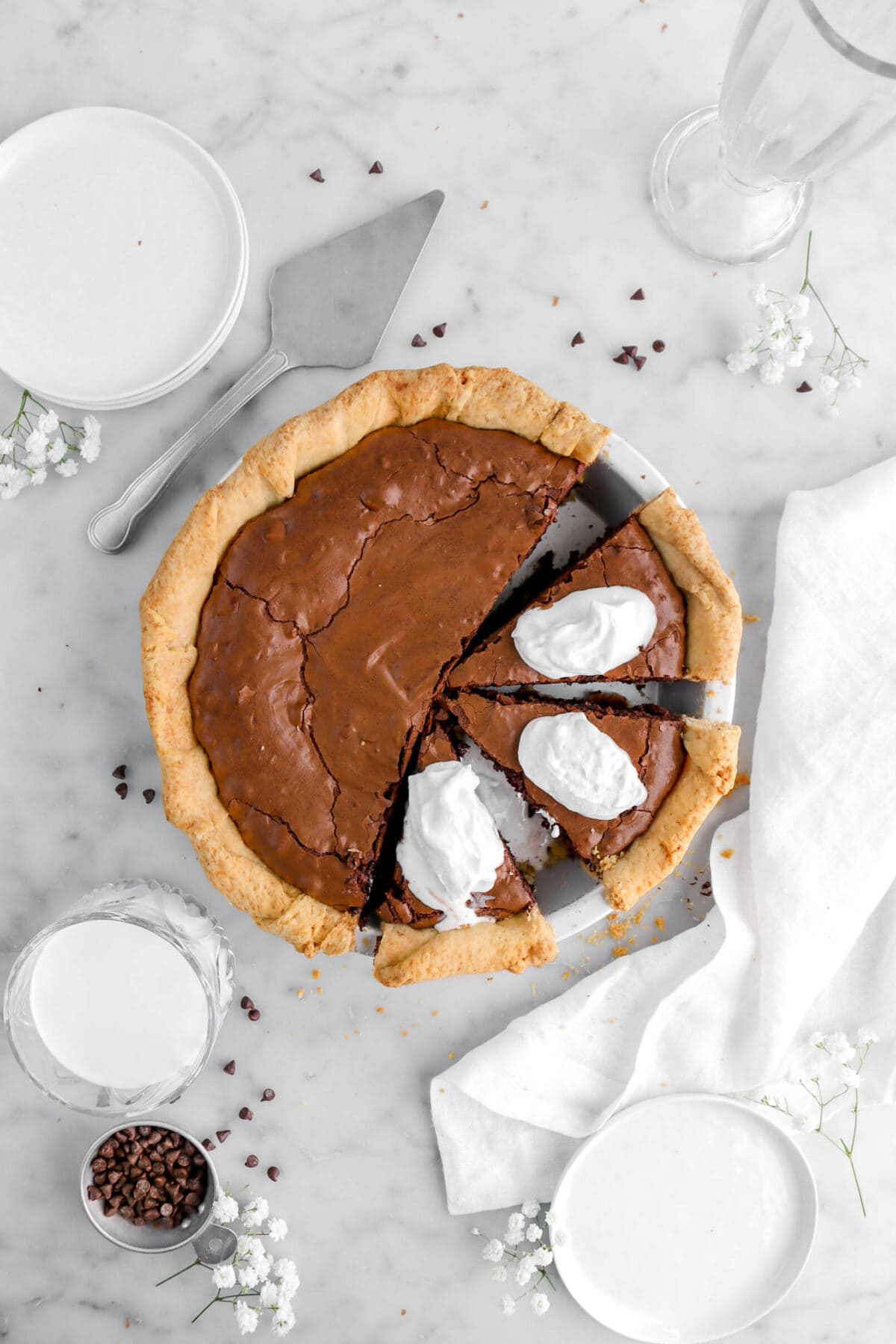 overhead shot of brownie pie with three slices cut into pie with all three slices having a dollop of whipped cream on top, a white napkin and flowers around, with chocolate chips and a glass of milk beside on marble surface.