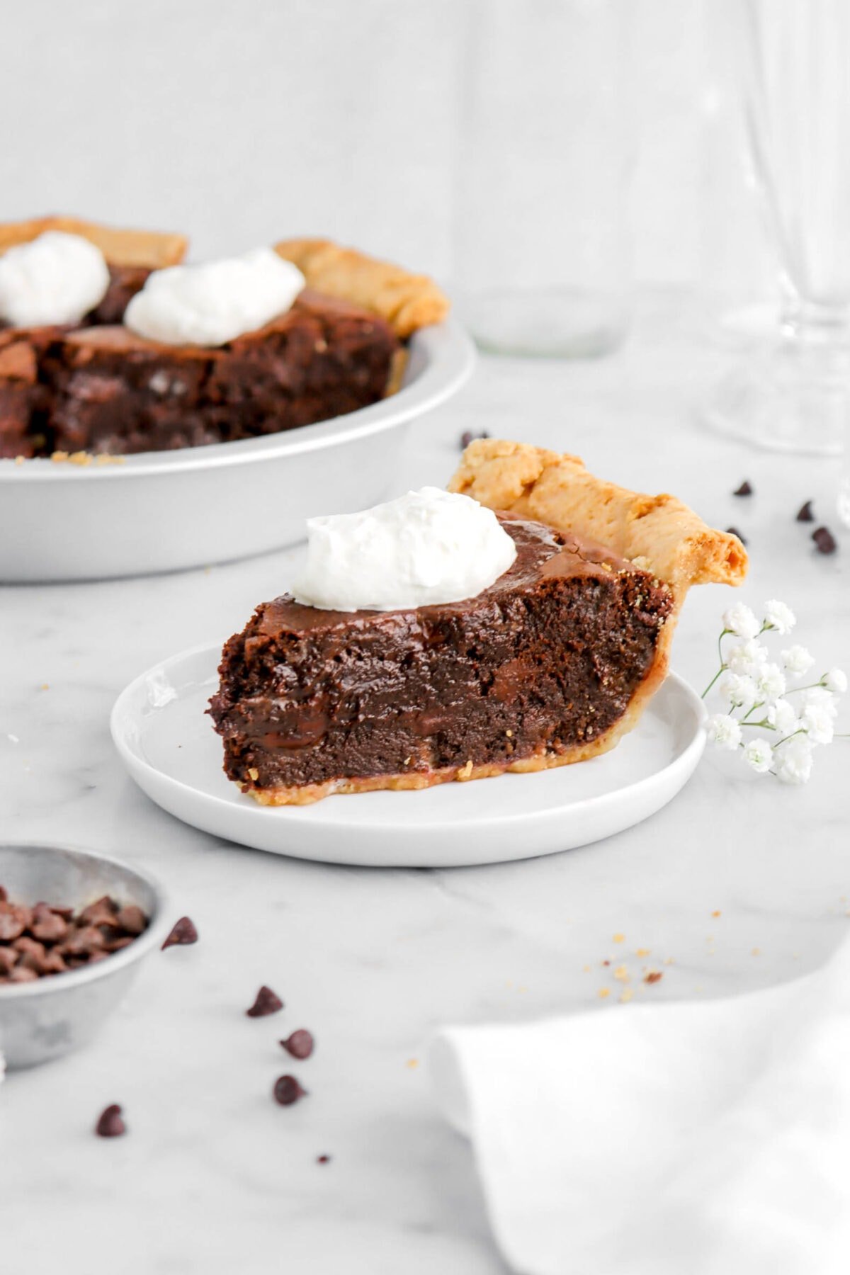 slice of brownie pie on white plate with full pie behind in metal pie plate on marble surface.