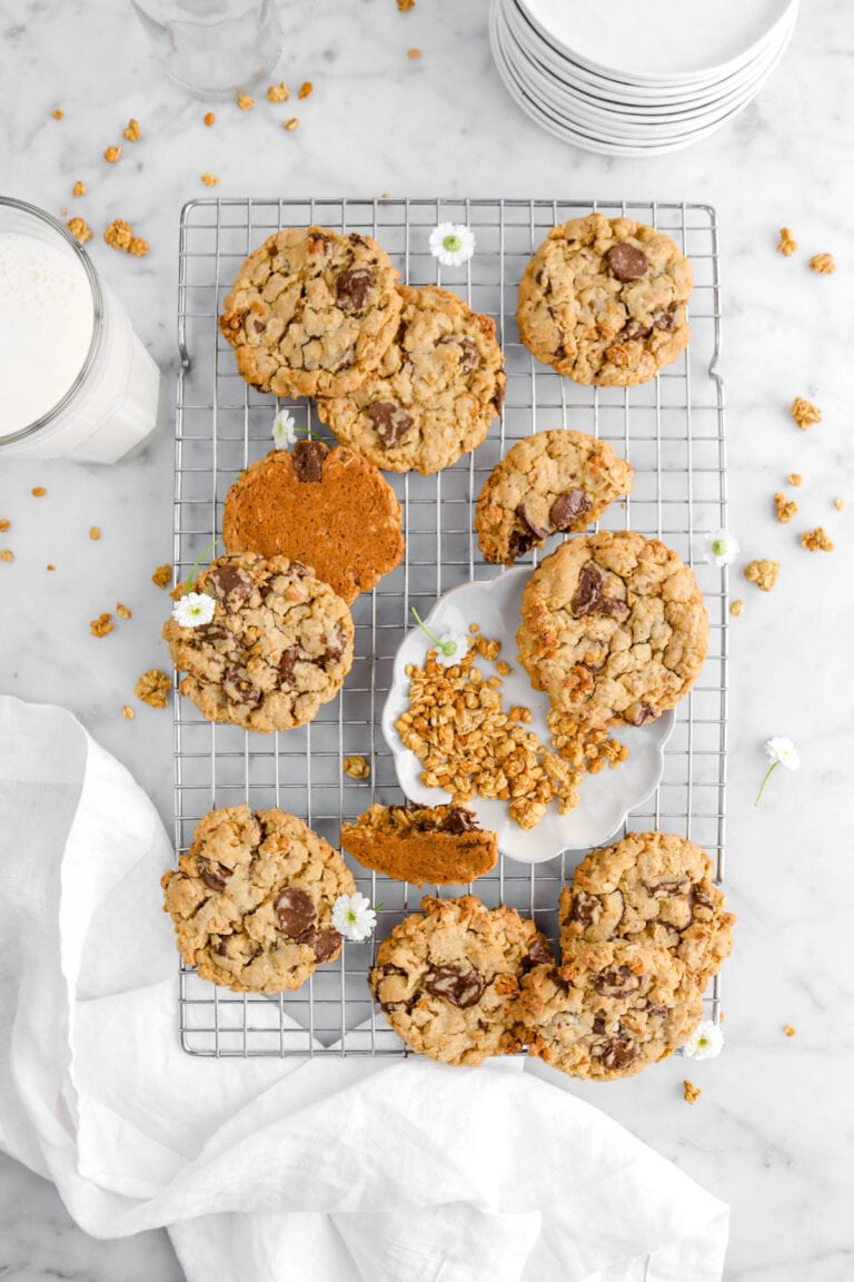 cookies on cooling rack with plate of granola and white flowers on cooling rack as well, a white napkin beside, a glass of milk, stack of plates, and granola scattered around on marble surface.