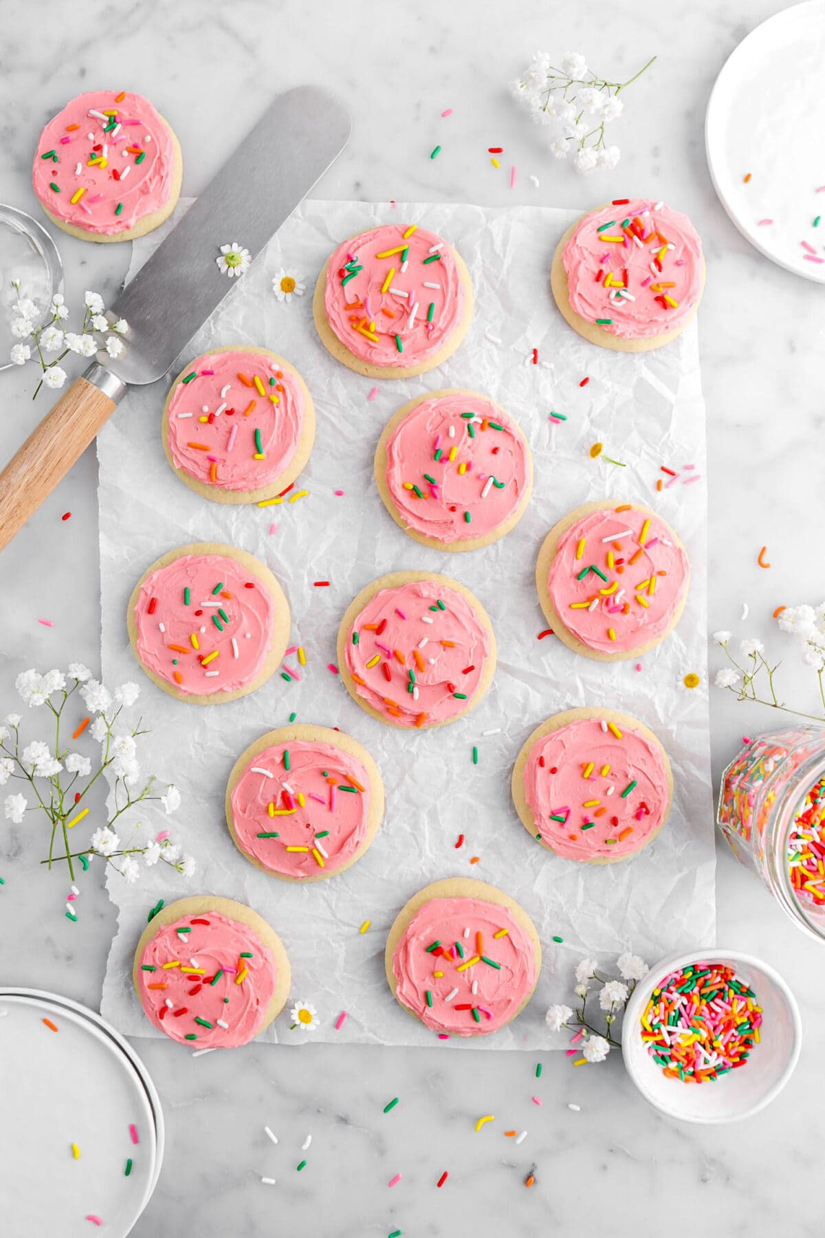 overhead shot of twelve frosted sugar cookies with flowers and rainbow sprinkles around, an offset spatula beside, white platees, a bowl, and a jar with sprinkles.