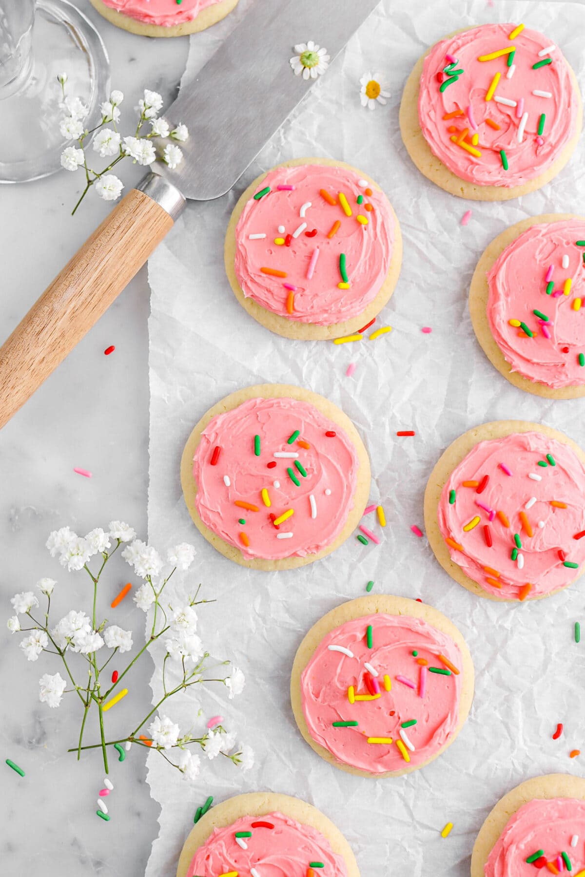 cropped overhead shot of eight frosted sugar cookies on parchment paper with flowers beside and an offset spatula.