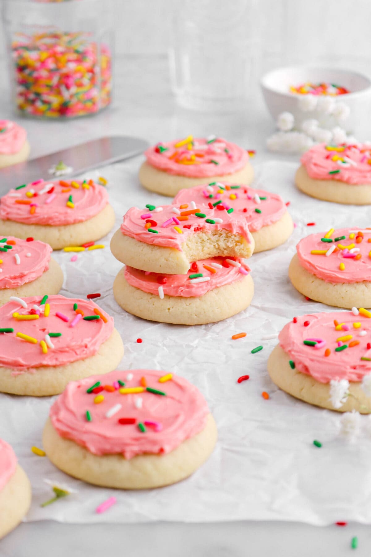 two stacked frosted sugar cookies on parchment paper with eleven cookies around, with white flowers and rainbow sprinkles around.