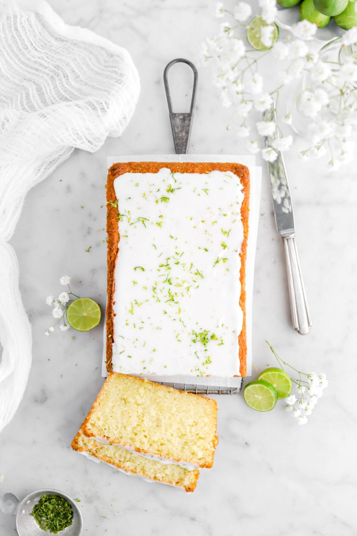 close up overhead shot of key lime pound cake with two slices laying in front with key limes around and white flowers, a white cheese cloth and a knife on either side of cake.