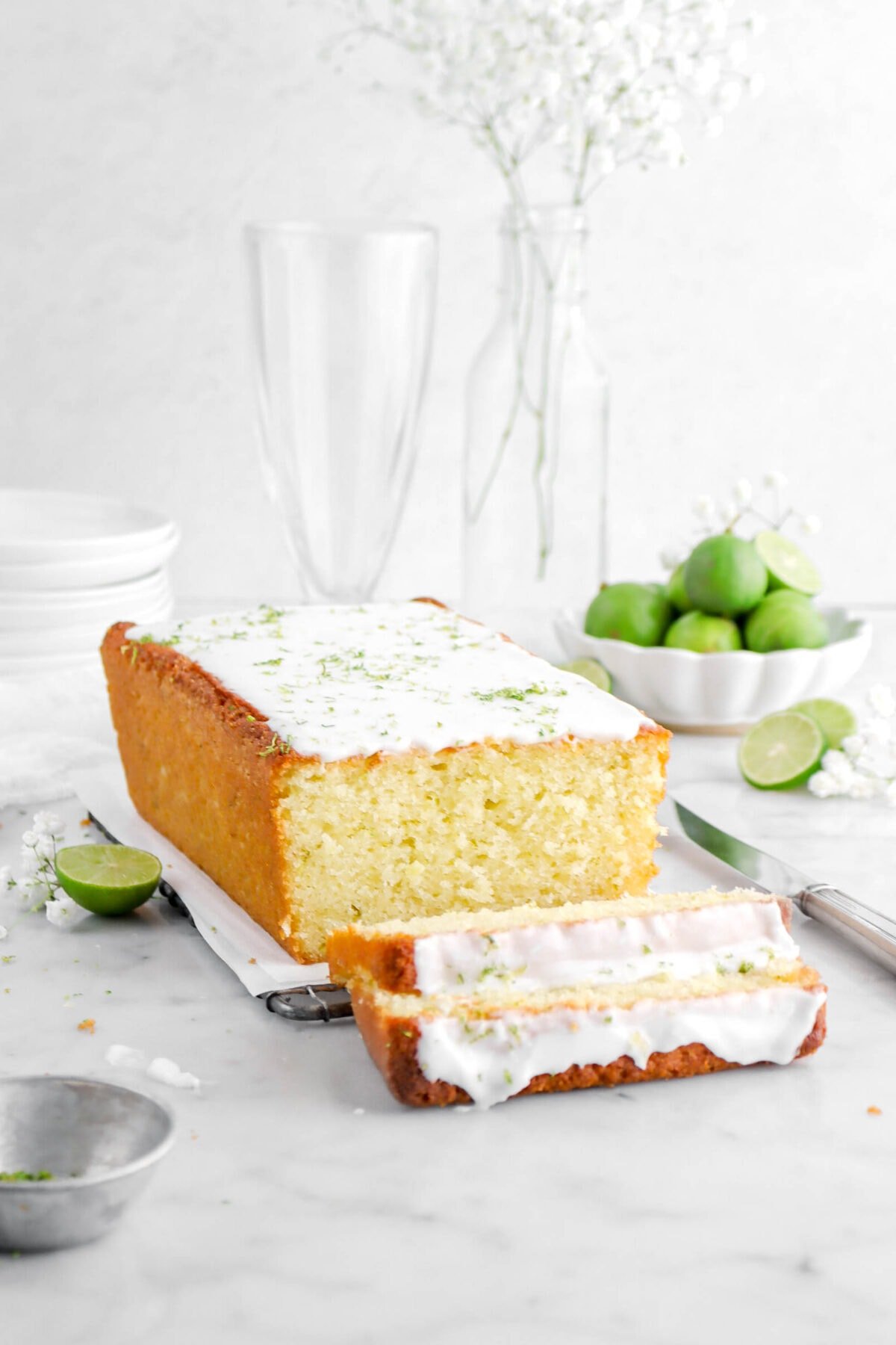 close up of key lime pound cake turned at an angle with two slices laying in front, key lies and white flowers around on marble surface.