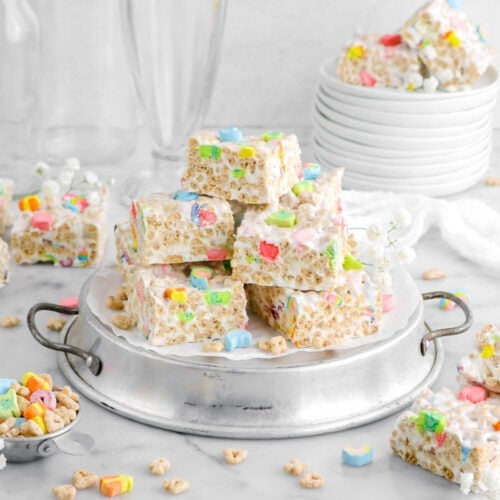 piled lucky charms marshmallow treats on upside down cake pan with stack of plates behind with more marshmallow treats and empty glasses behind with more cereal scattered around.
