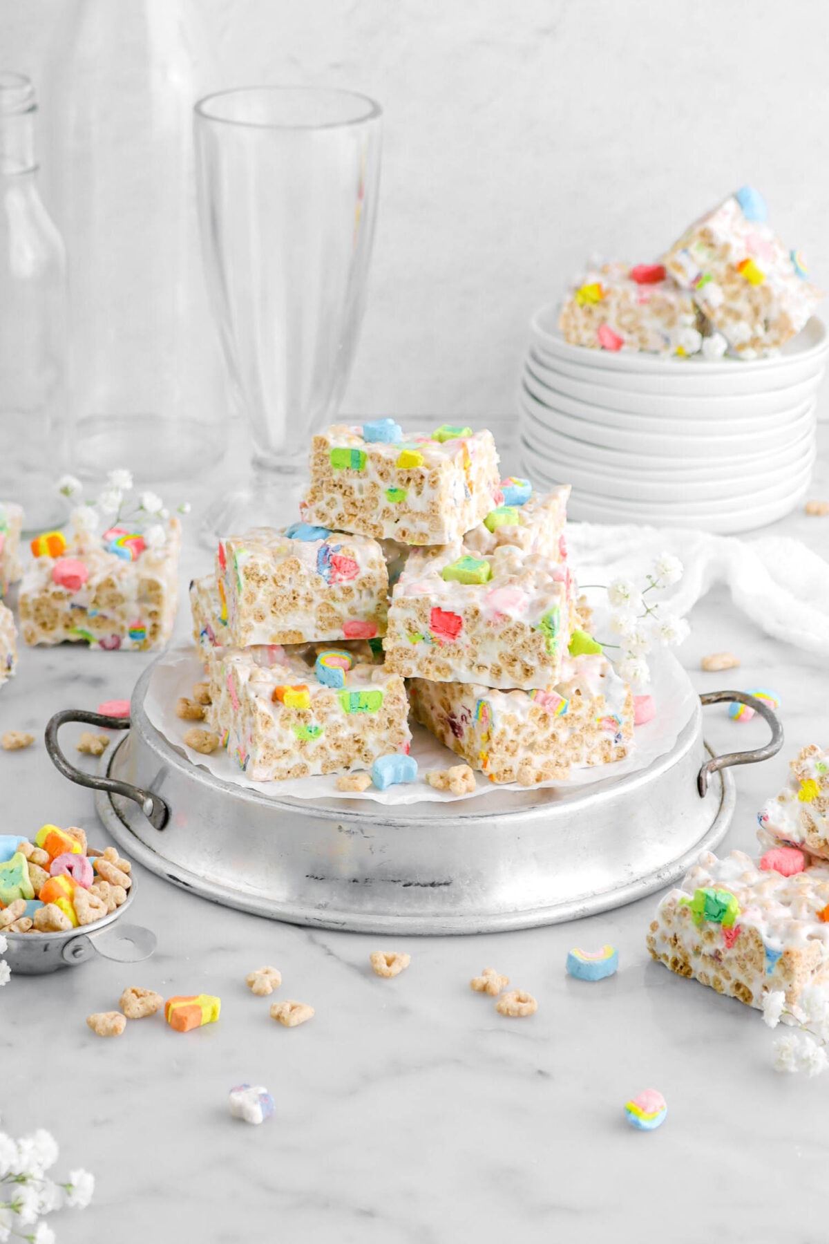 piled lucky charms marshmallow treats on upside down cake pan with stack of plates behind with more marshmallow treats and empty glasses behind with more cereal scattered around.