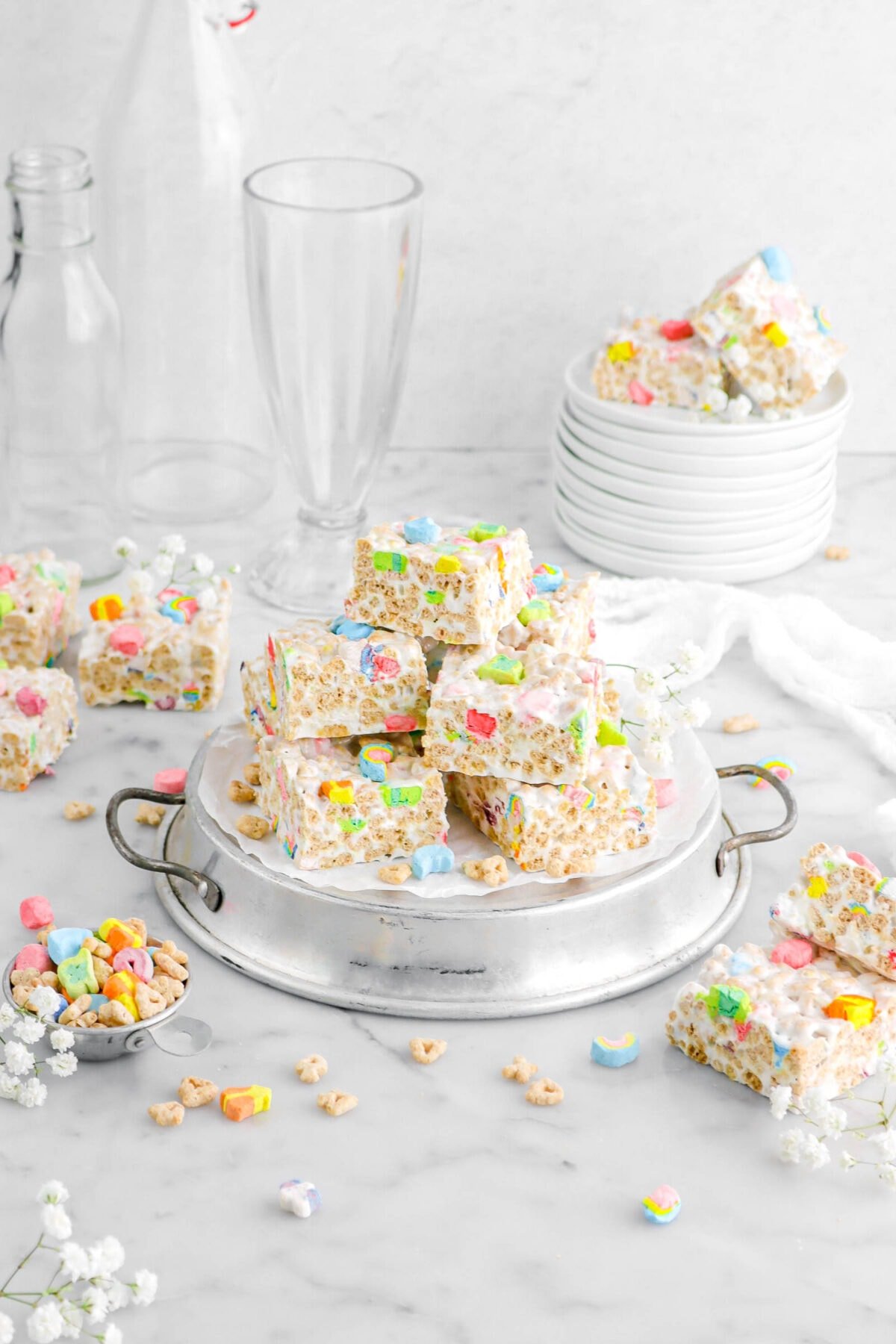 pulled back shot of lucky charms marshmallow treats stacked on upside down cake pan with measuring cup of cereal beside, more marshmallow treats around, a stack of white plates, white flowers, and empty glasses behind.