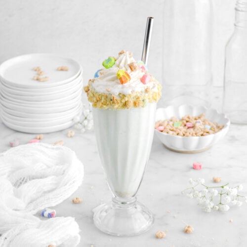 angled shot of milkshake in tall glass with cereal on top and whipped cream with a white cheese cloth beside, a stack of white plates behind, and more cereal around.