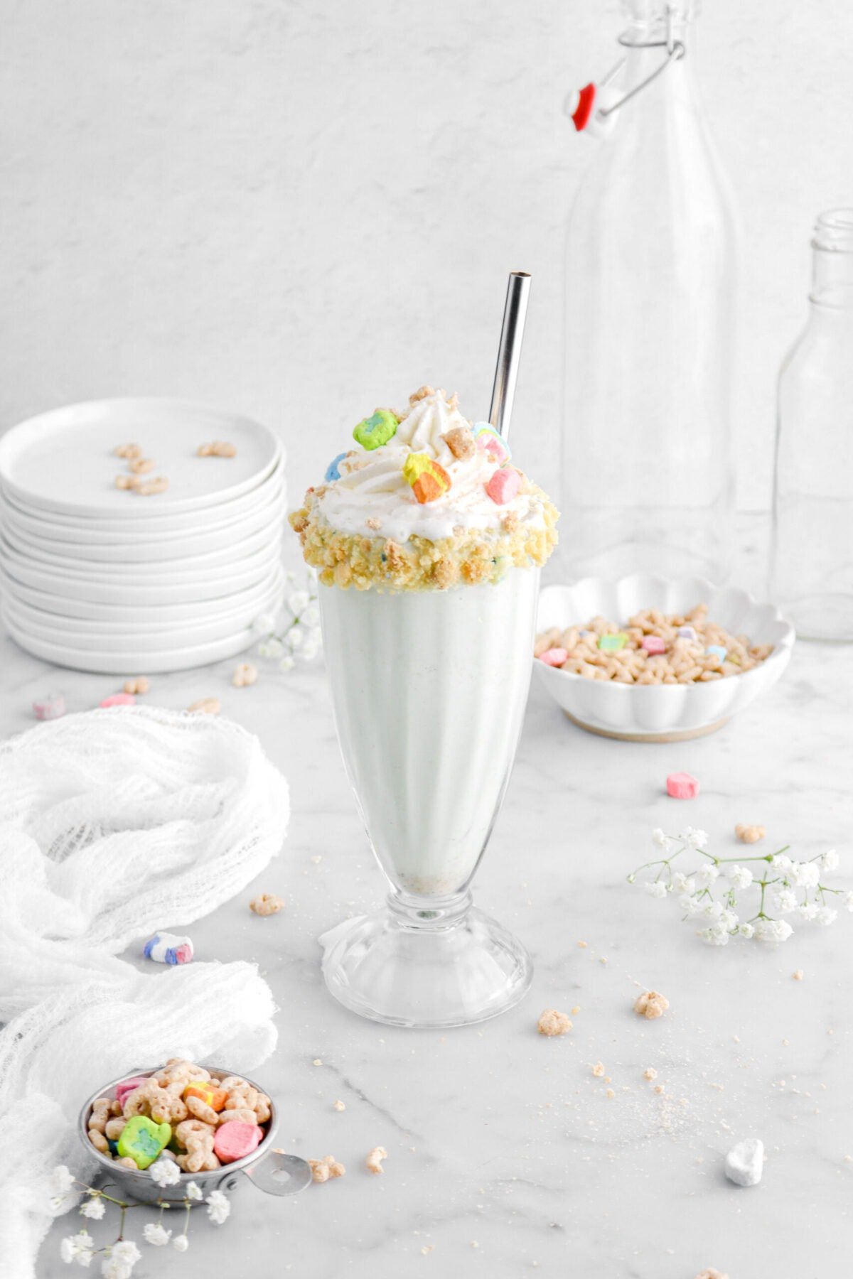 angled shot of milkshake in tall glass with cereal on top and whipped cream with a white cheese cloth beside, a stack of white plates behind, and more cereal around.