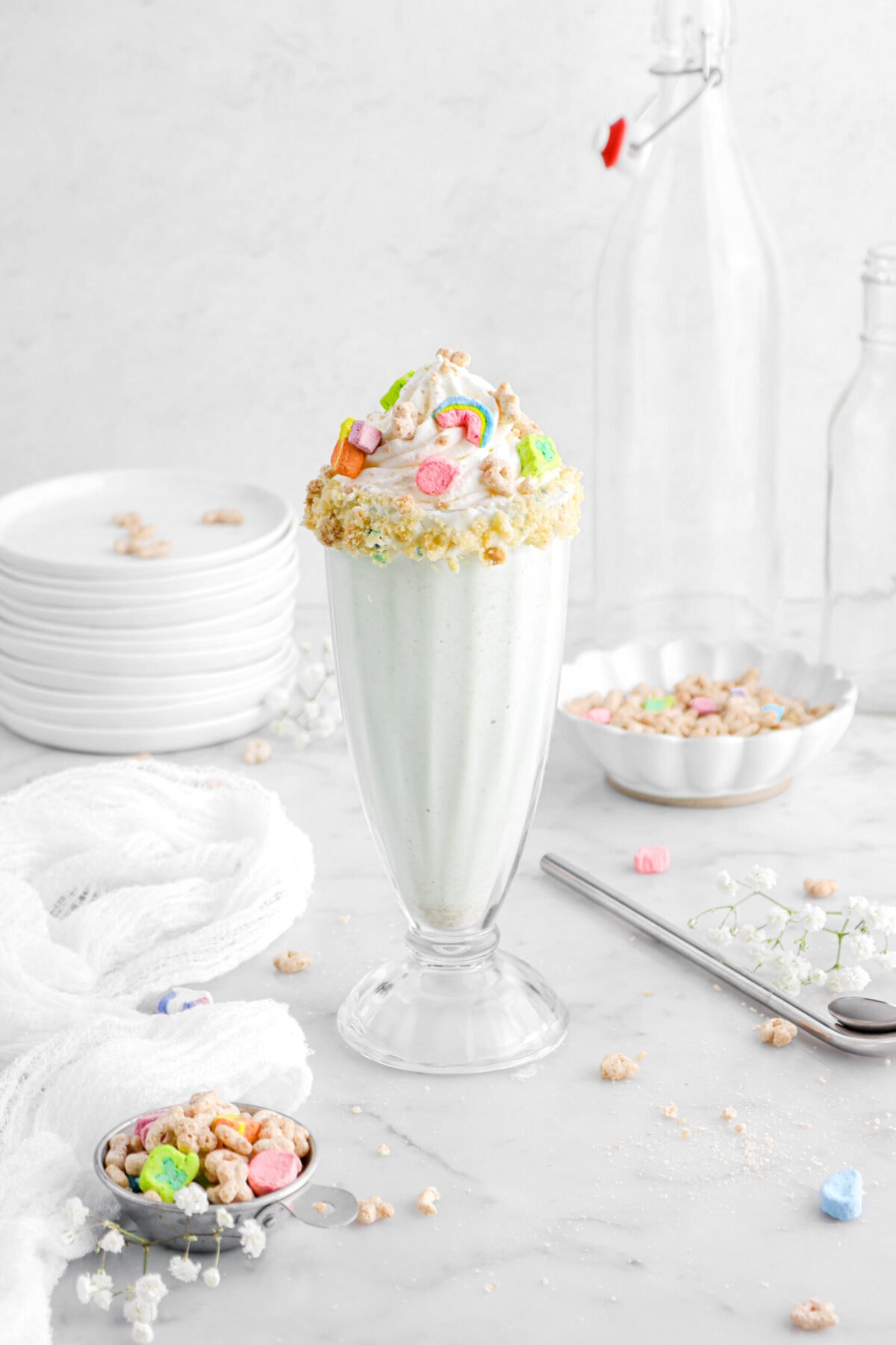 lucky charms milkshake in tall milkshake glass with a stack of plates, two empty glasses, and a scalloped bowl with cereal behind, a metal straw beside with a white cheese cloth.