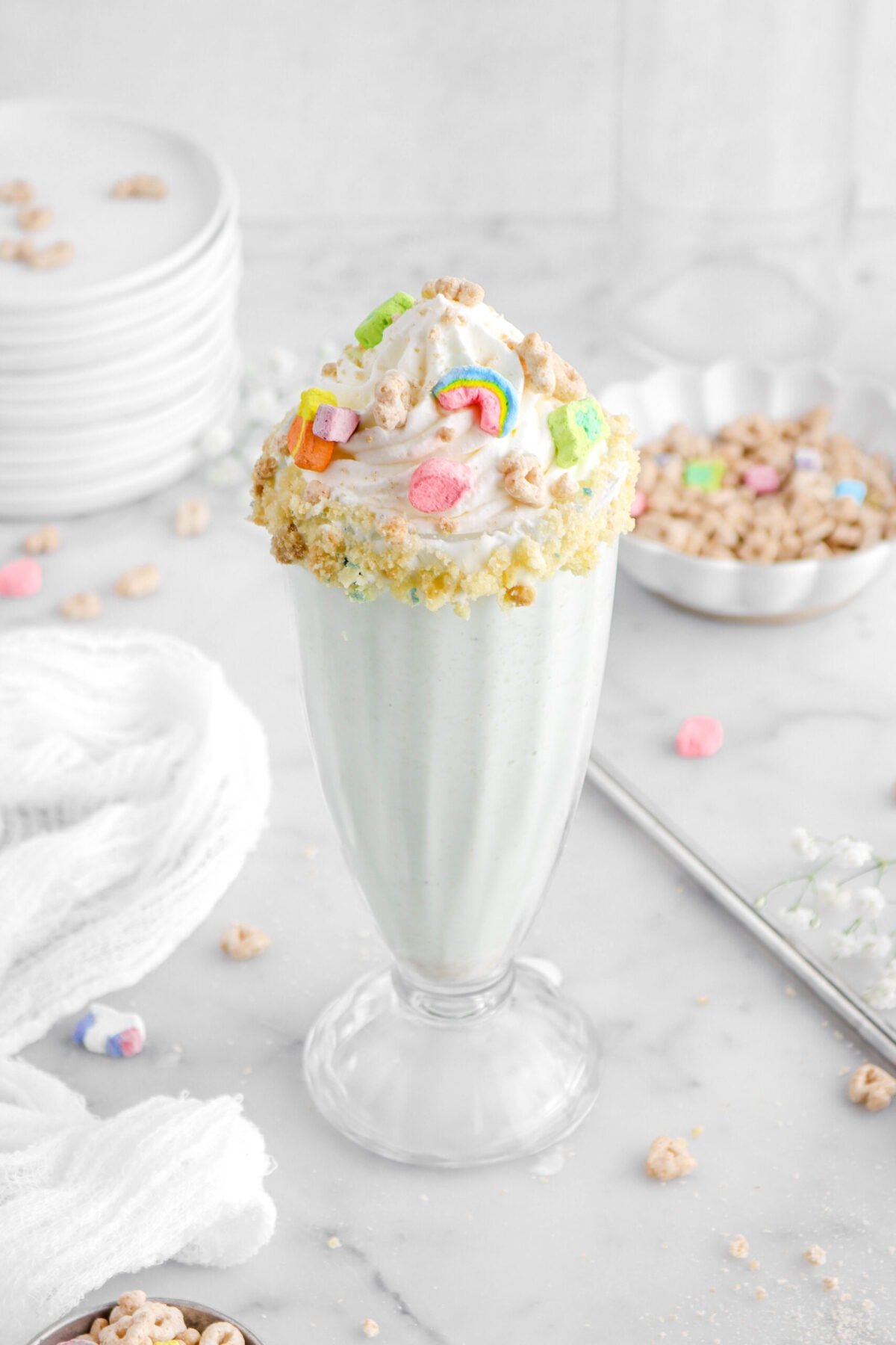 angled close up of lucky charms milkshake in tall glass with whipped cream and cereal on top, with a bowl of cereal behind and a stack of plates with white flowers around.