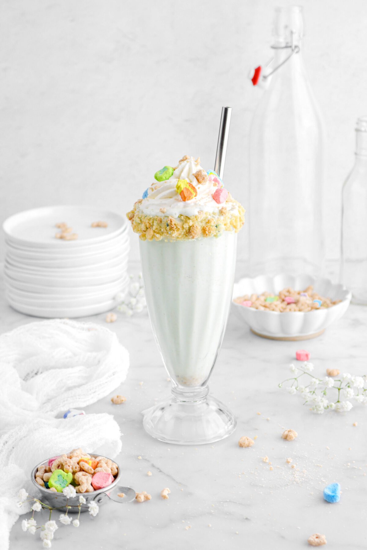 lucky charms in milkshake glass with metal straw, more cereal around, and white flowers.