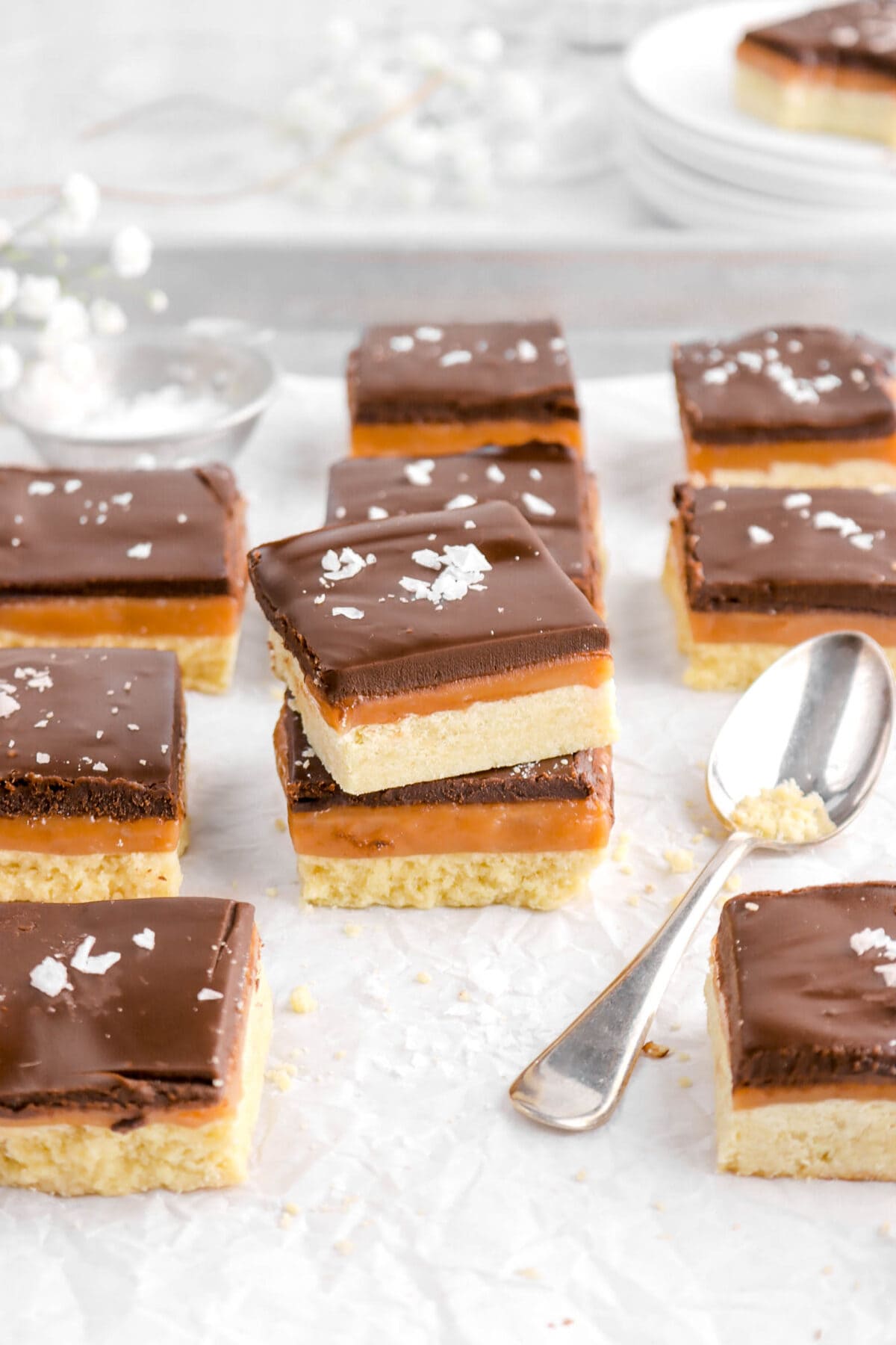 angled overhead shot of millionaire's shortbread bars in sheet pan with spoon beside, measuring cup of salt, and white flowers behind.