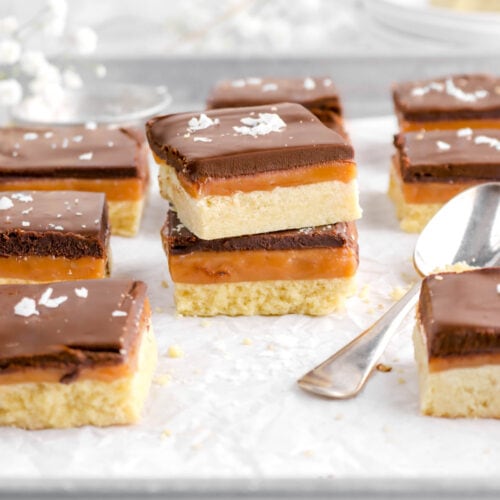 front shot of two stacked millionaire's shortbread bars seven around on sheet pan with spoon beside and white flowers behind, as well as stack of plates and another bar with bite missing.