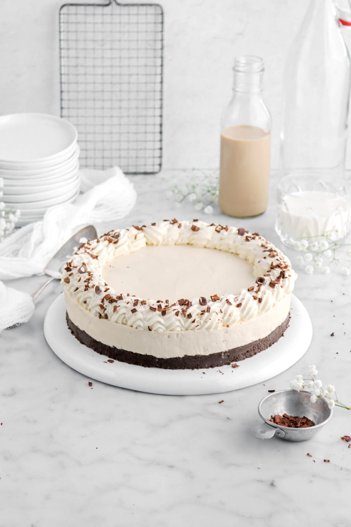 irish cream cheesecake on upside down plate with glass of milk and irish cream behind, stack of plates, and white flowers aorund.