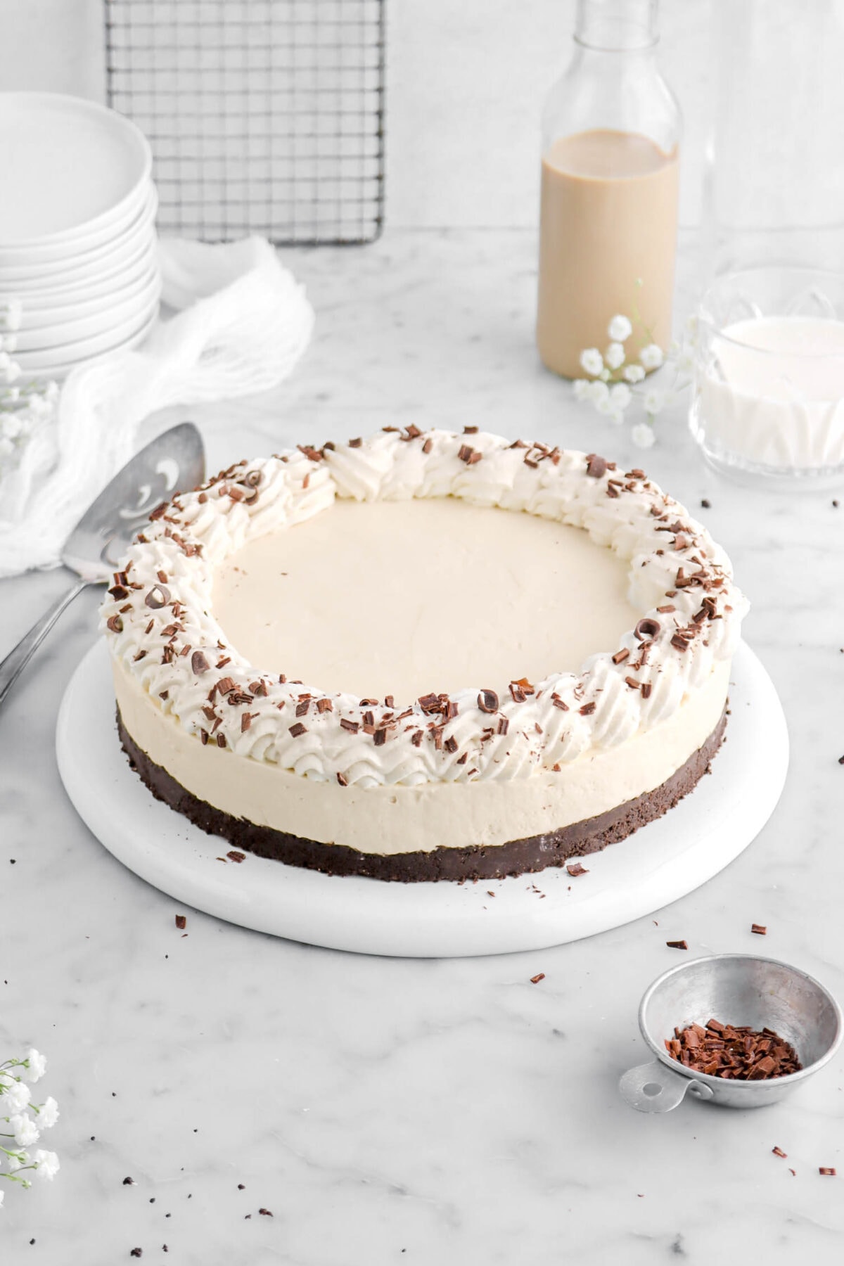 angled shot of irish cream cheesecake on upside down white palte with a cake knife, measuring cup of chocolate curls, and white flowers around on marble surface.