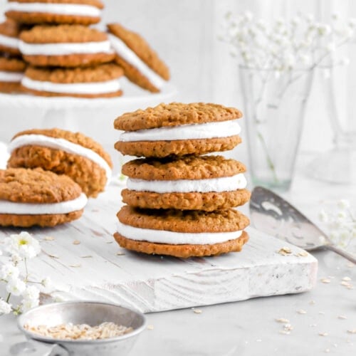 three stacked oatmeal cream pies on white wood board with two ore behind and a pile of them on white cake plate behind.