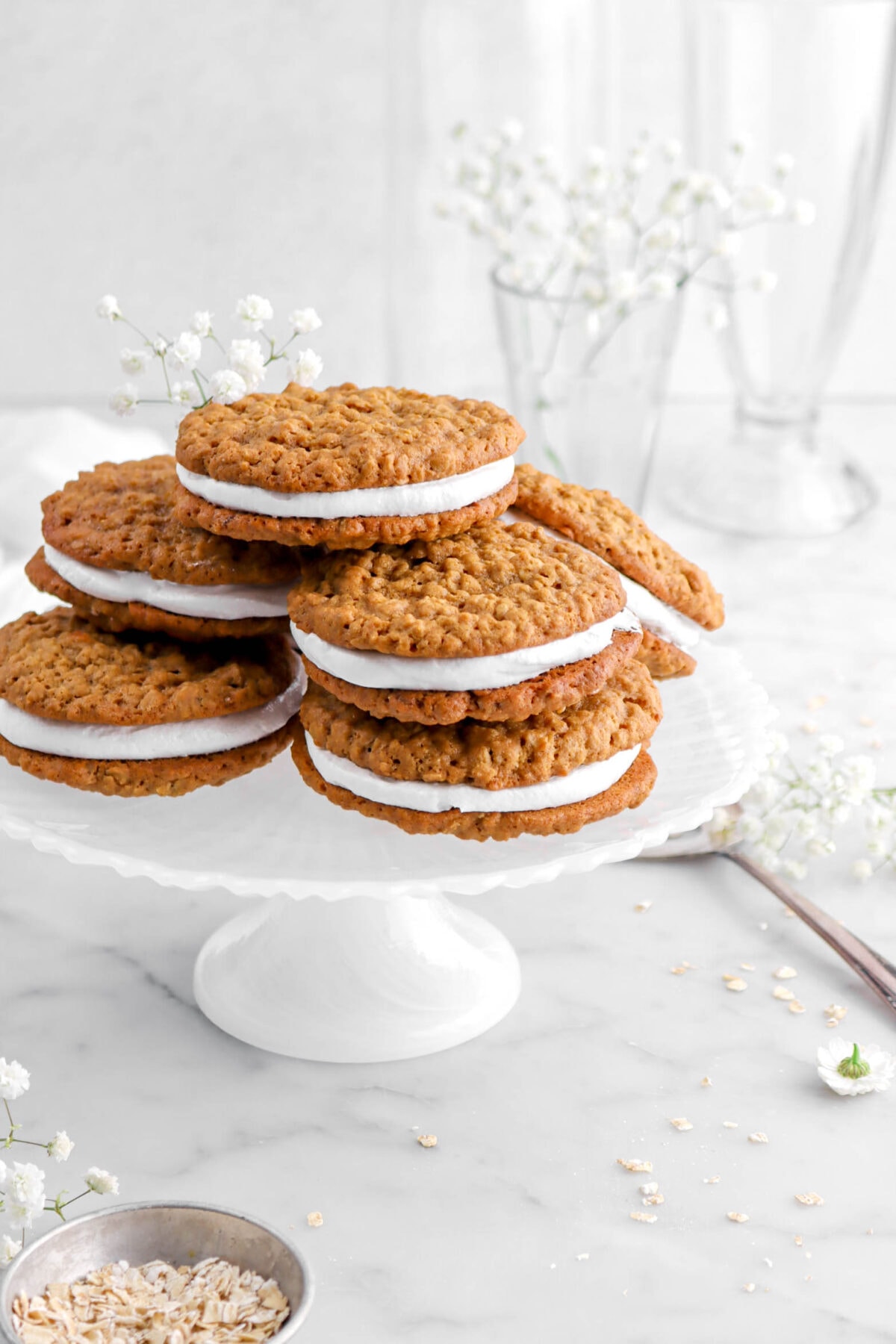 cropped angled shot of oatmeal cream pies on white cake plate with flowers on top and flowers around on marble surface with a measuring up of oatmeal beside.