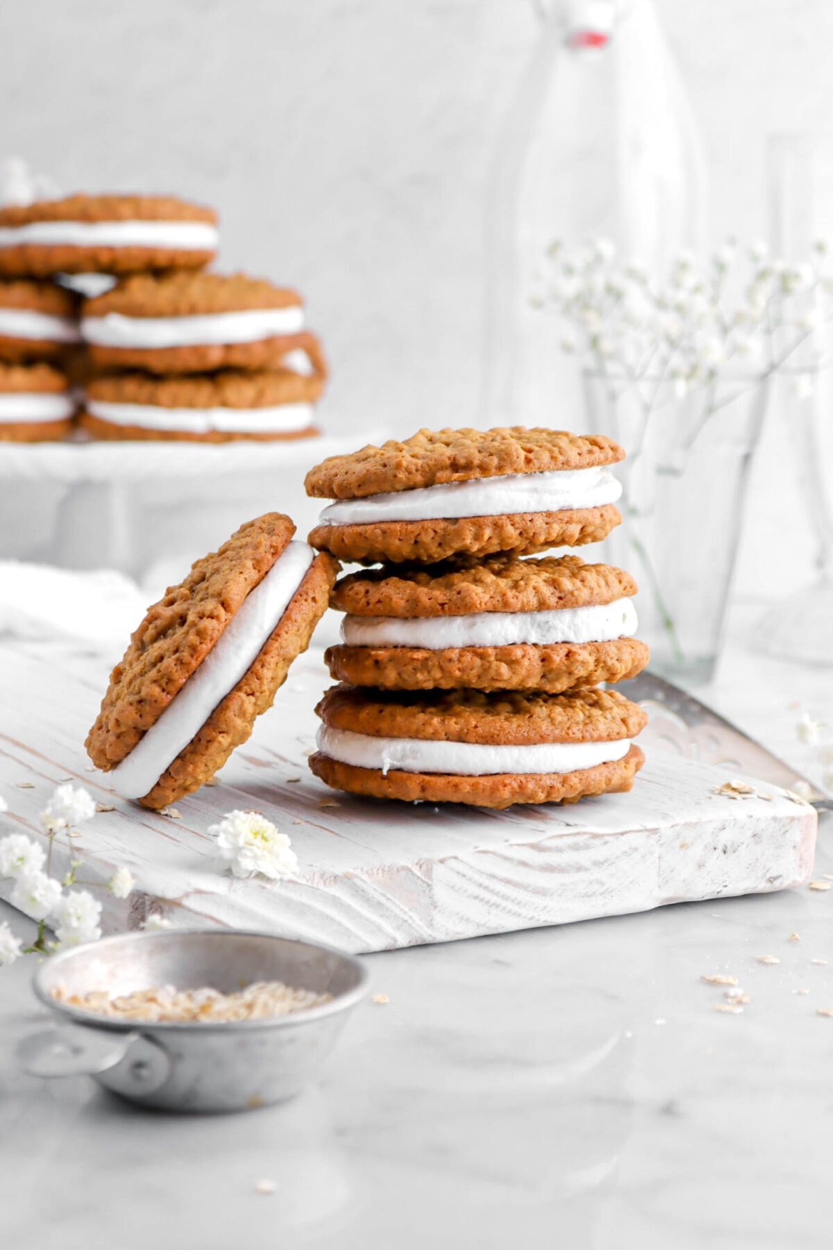 three stacked oatmeal cream pies with one leaning against stack on wood board with more oatmeal cream pies behind on white cake plate.