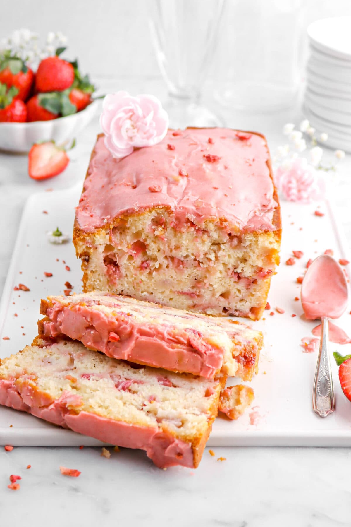angled close up of strawberry bread with two slices laying in front on white tray with a pink carnation on top and a spoon of strawberry icing beside.