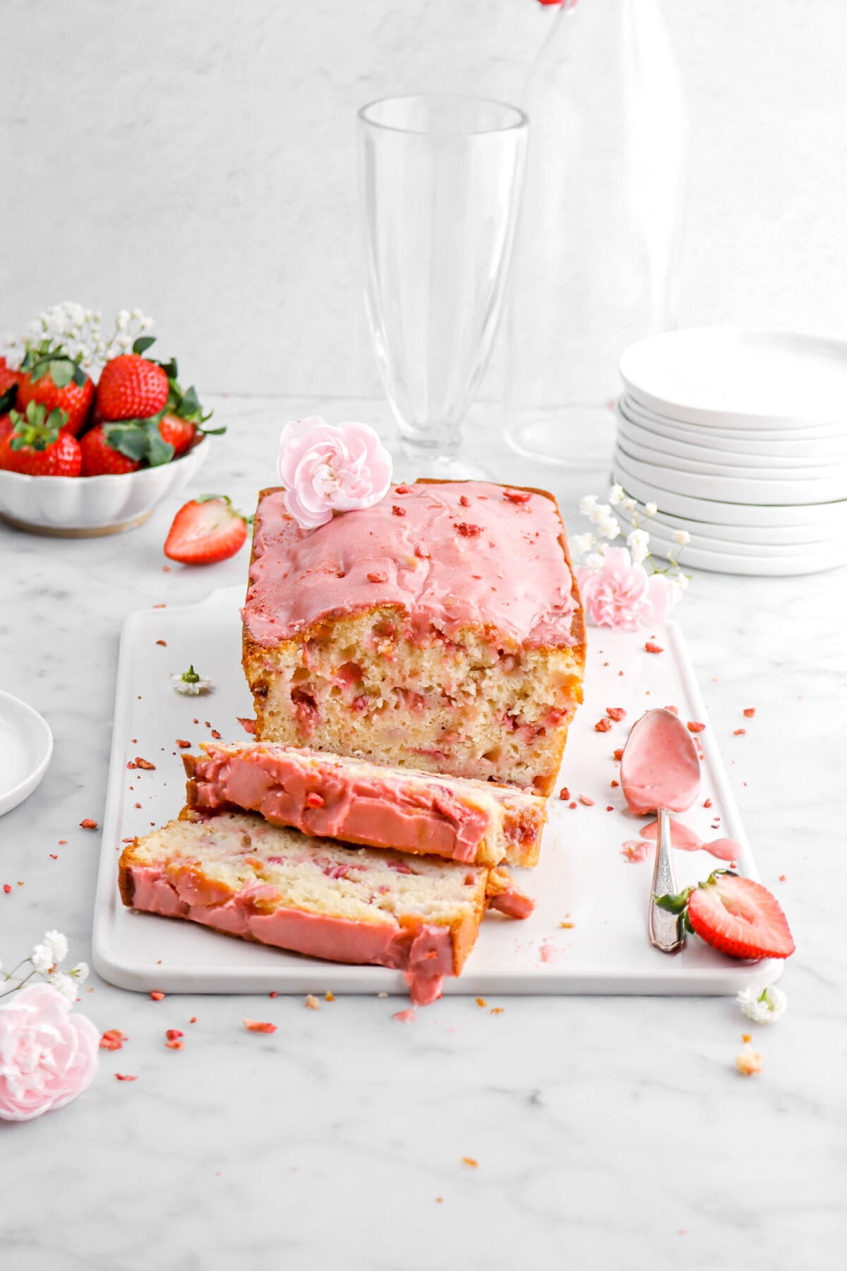 strawberry bread on white tray with two slices laying in front with flowers around and fresh strawberries, a stack of plates and two empty glasses behind.