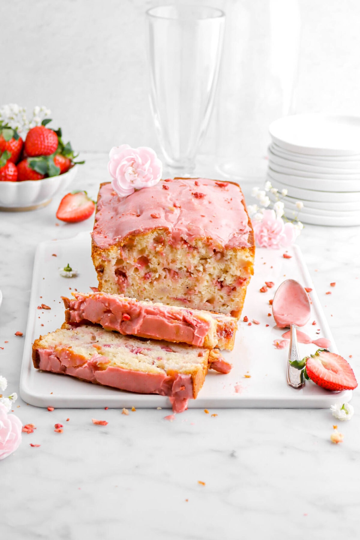 strawberry bread with strawberry icing and pink carnation on top, two slicing laying in front on white tray with fresh strawberries around on marble surface.