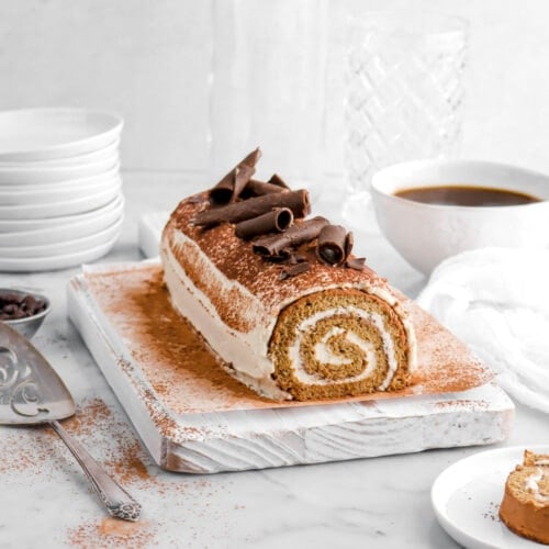 tiramisu cake roll on white board with cocoa powder sprinkled around, a cake knife and measuring cup of coffee beans beside, a stack of plates, two empty glasses, and a white mug of coffee behind.