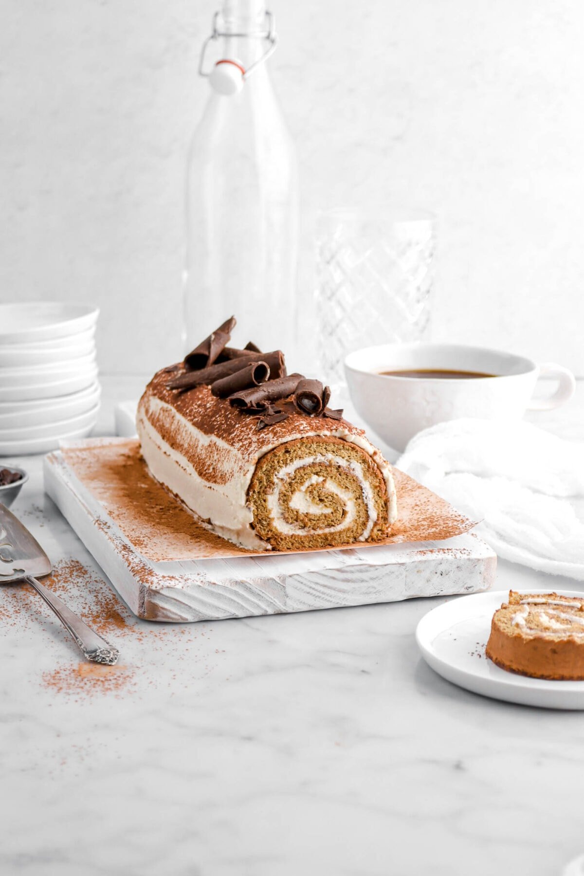 front shot of tiramisu cake roll on white wood board with cocoa powder and chocolate curls on top, a white plate with a slice of cake roll in front, a coffee mug behind with a white cheesecloth and two empty glasses.