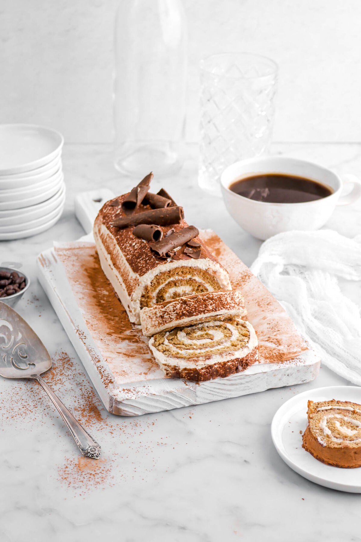two slices of cake in front of tiramisu cake roll on white wood board with another slice of cake on white plate in front, a cup of coffee behind, and a cake knife beside.