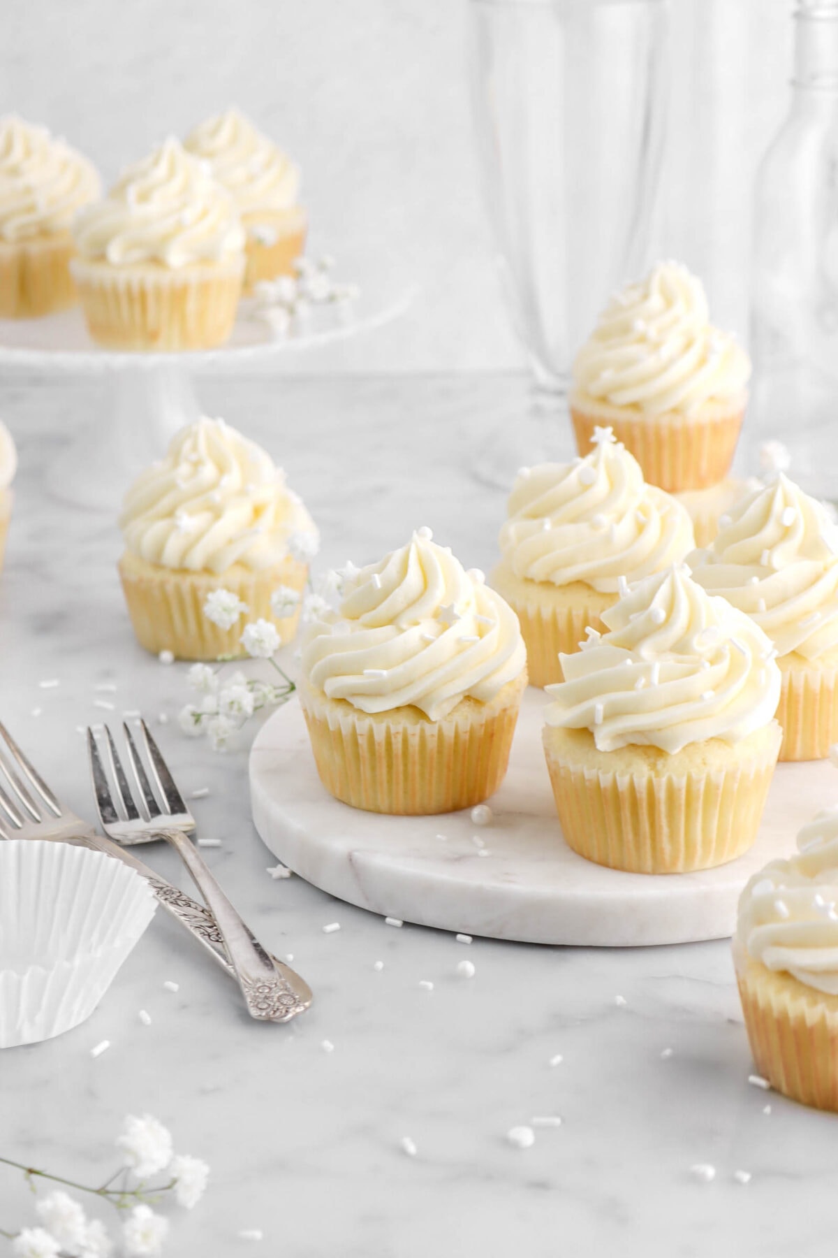 four vanilla cupcakes on marble plate with more cupcakes around with white sprinkles around, white flowers, two forks, and an empty cupcake paper beside.
