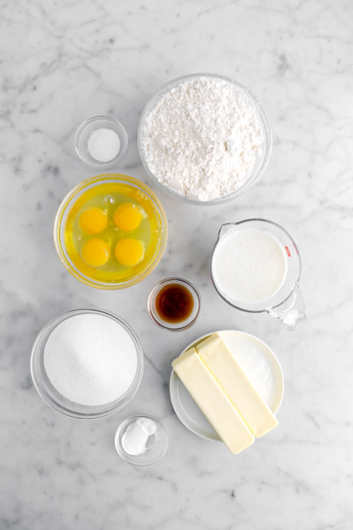 salt, flour, eggs, vanilla, milk, sugar, butter, and baking powder on marble surface.
