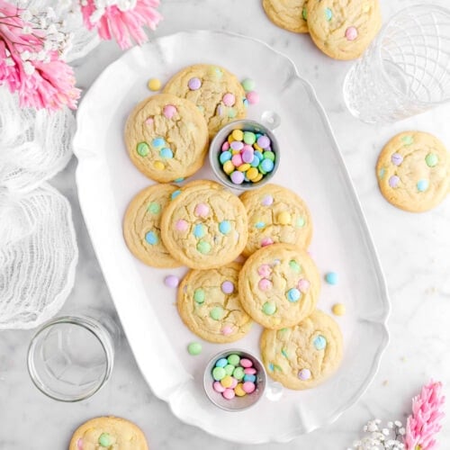 cookies on oval platter with two measuring cups of pastel M&M's, with more cookies around, two empty glasses, and a white cheesecloth on marble surface.