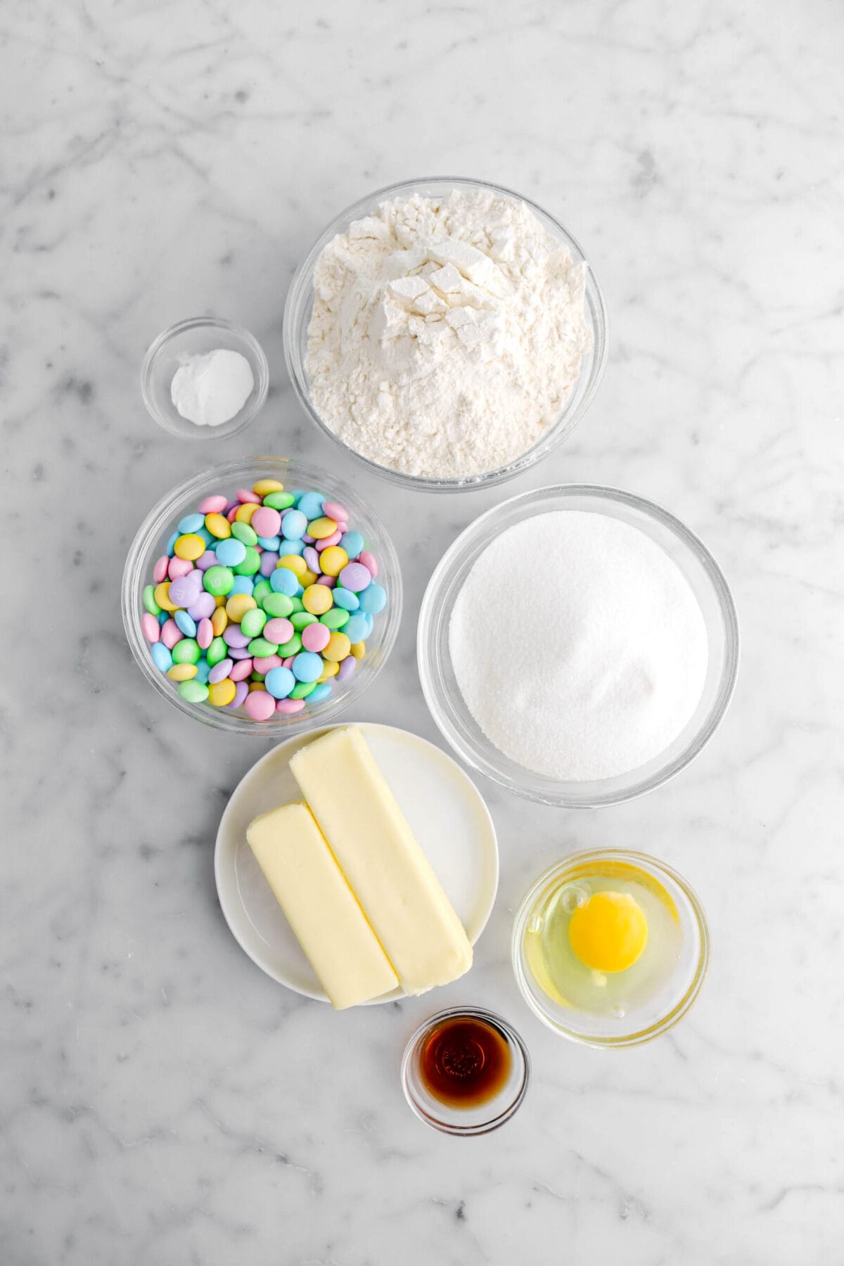 baking soda, flour, pastel M&M's, sugar, butter, egg, and vanilla on marble surface.