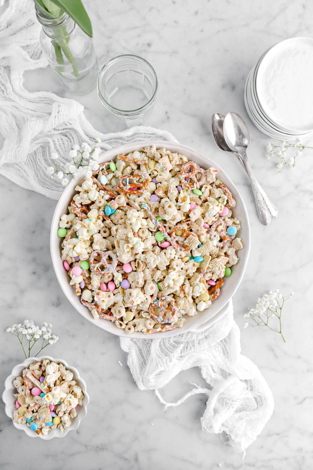 easter snack mix in white casserole on top of white cheesecloth with white flowers around, two spoons, and stack of white plates beside.