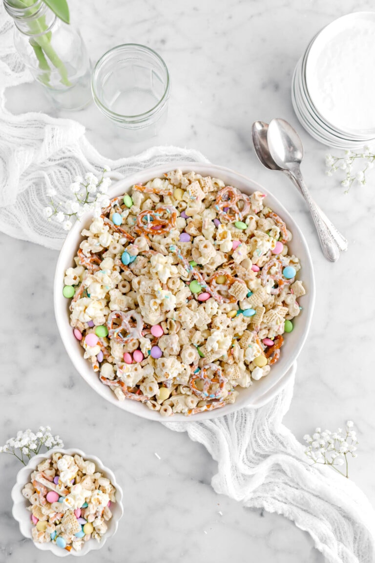 easter snack mix in round white casserole white white flowers around, a small bowl of snack mix beside, with spoons, empty jar, and stack of white paltes above.