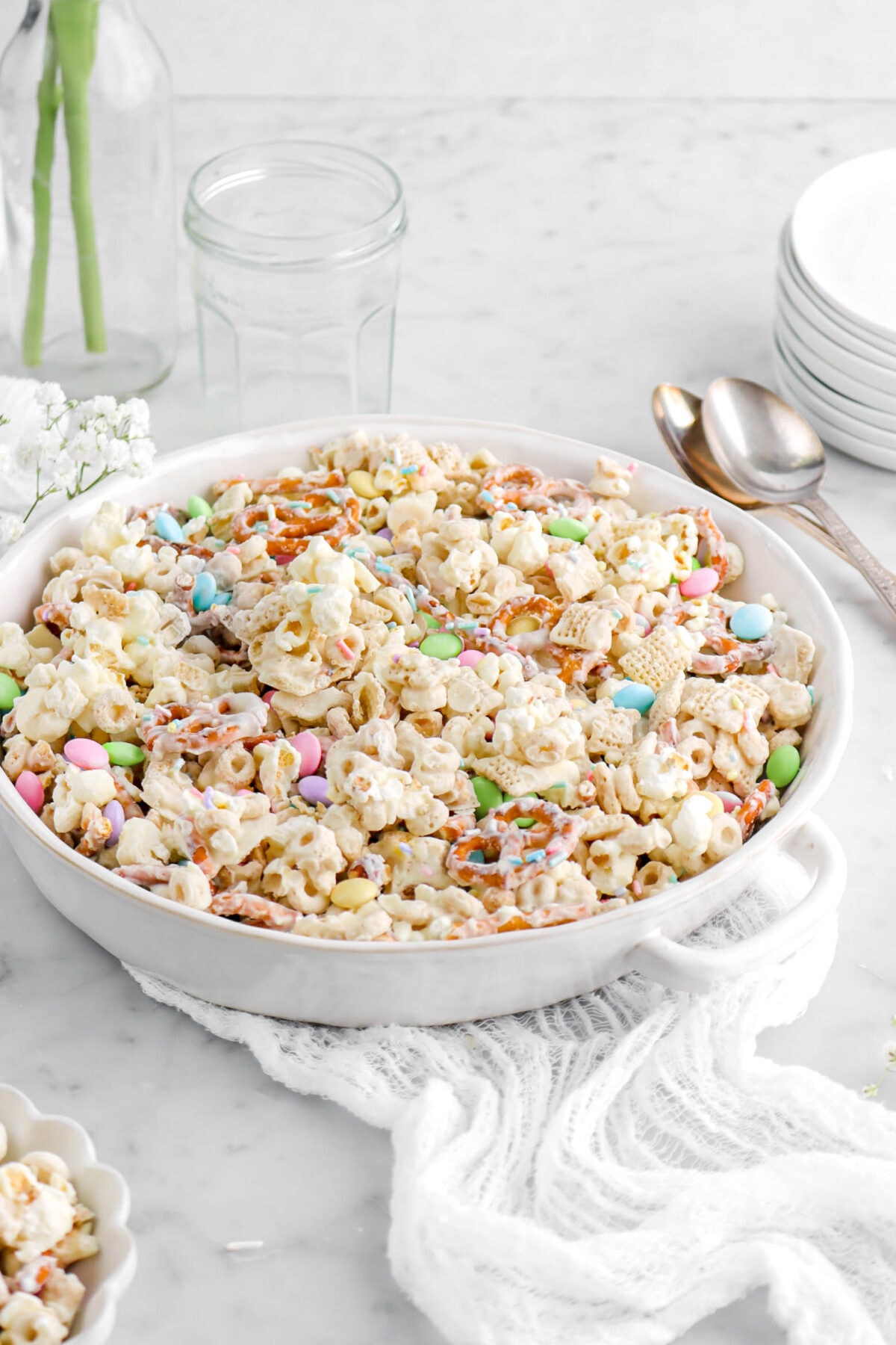 cropped angled photo of easter snack mix in round casserole with two spoons, empty jar, and stack of white plates behind.