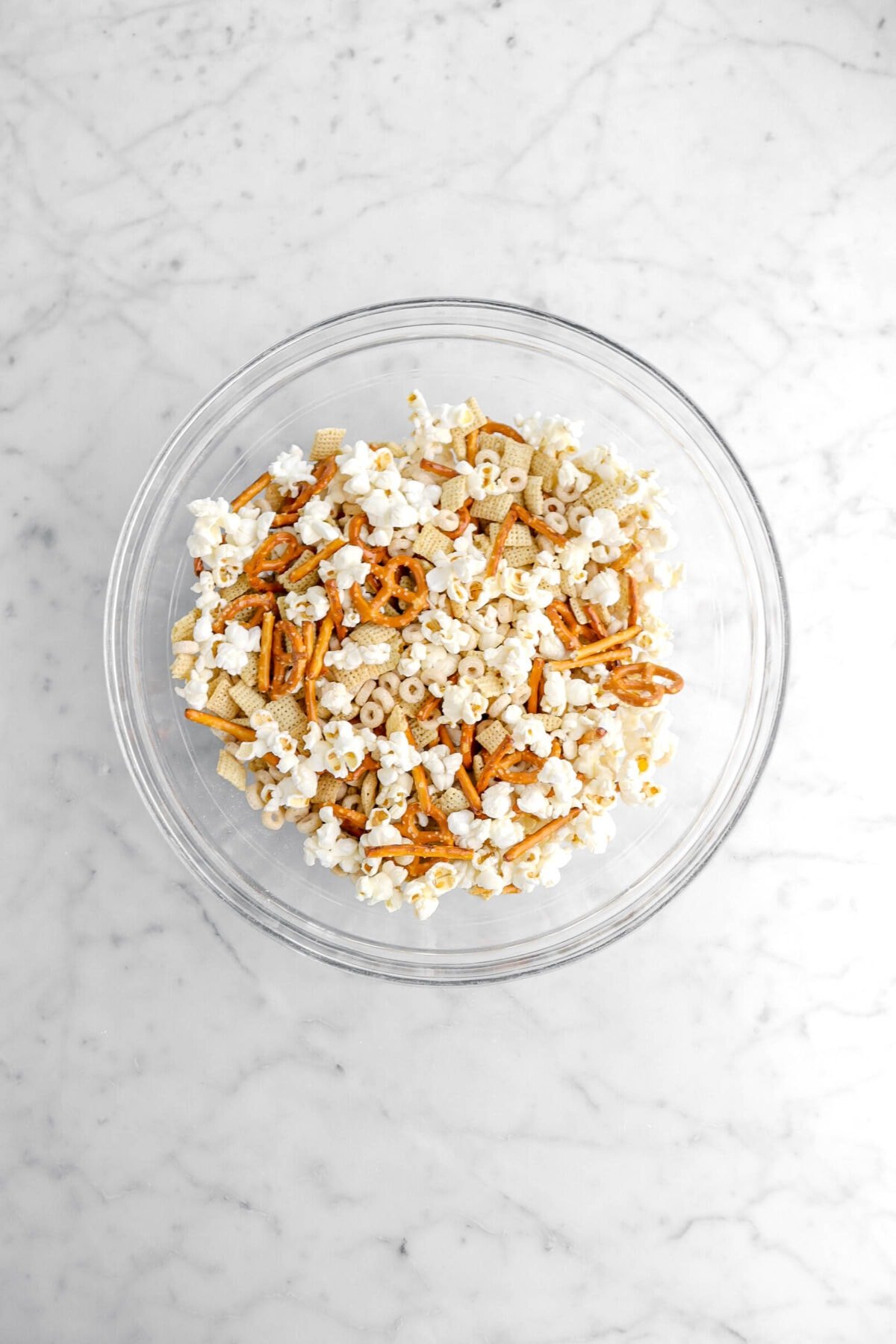 popcorn, pretzels, chex, cheerios, and peanuts mixed together in glass bowl.